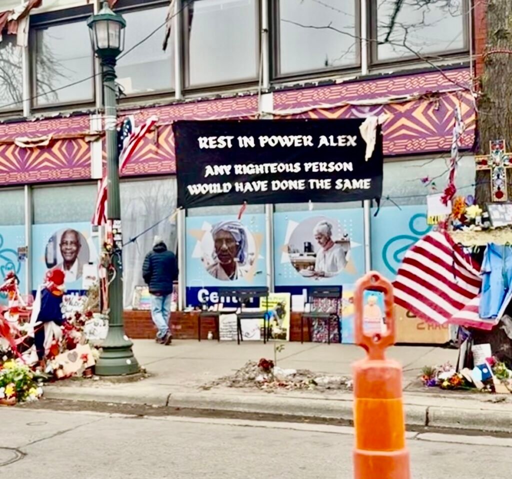 A photo of the street where Alex Pretti was murdered by ICE, now lined with memorials dedicated to him, including a banner stating "REST IN POWER ALEX - ANY RIGHTEOUS PERSON WOULD HAVE DONE THE SAME"